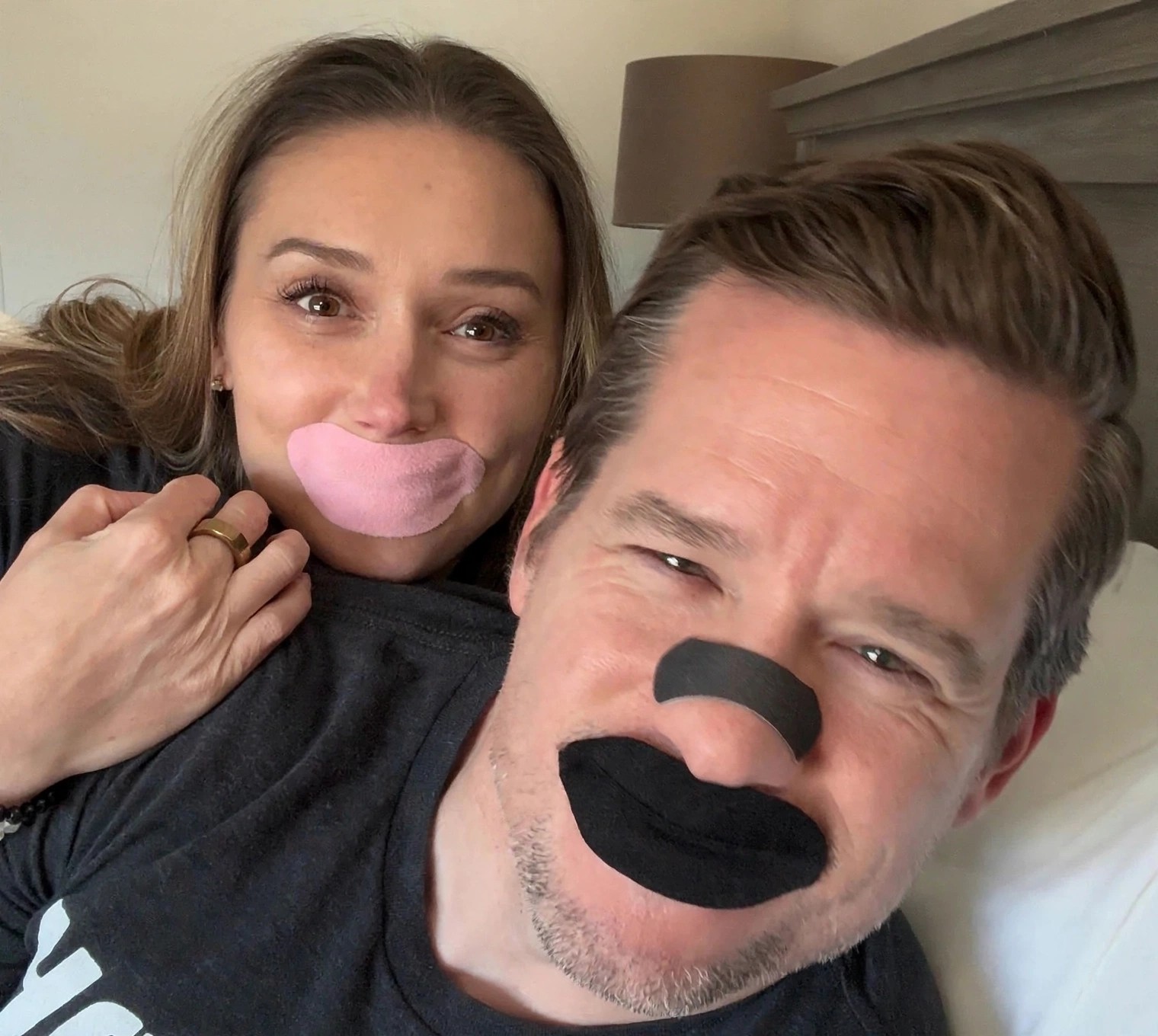 Couple in bed wearing pink and black mouth tape.